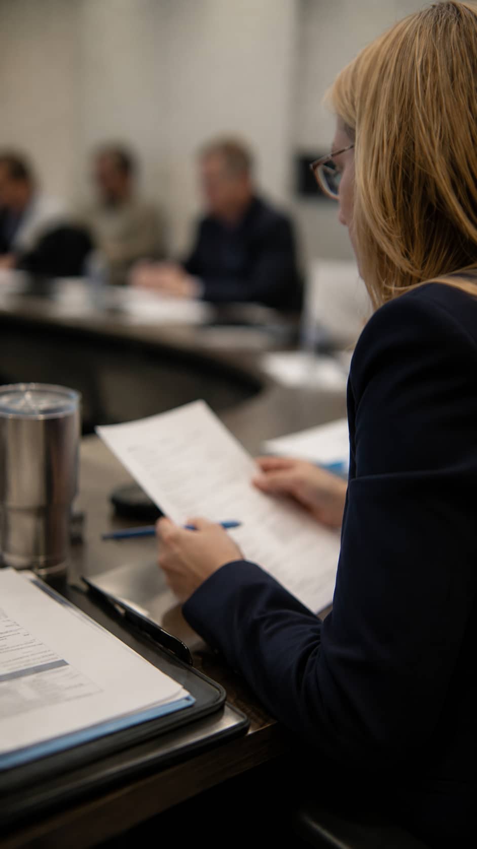 A woman in a dark blazer reviews a printed meeting document with a pen while seated at a boardroom table, with other attendees blurred in the background