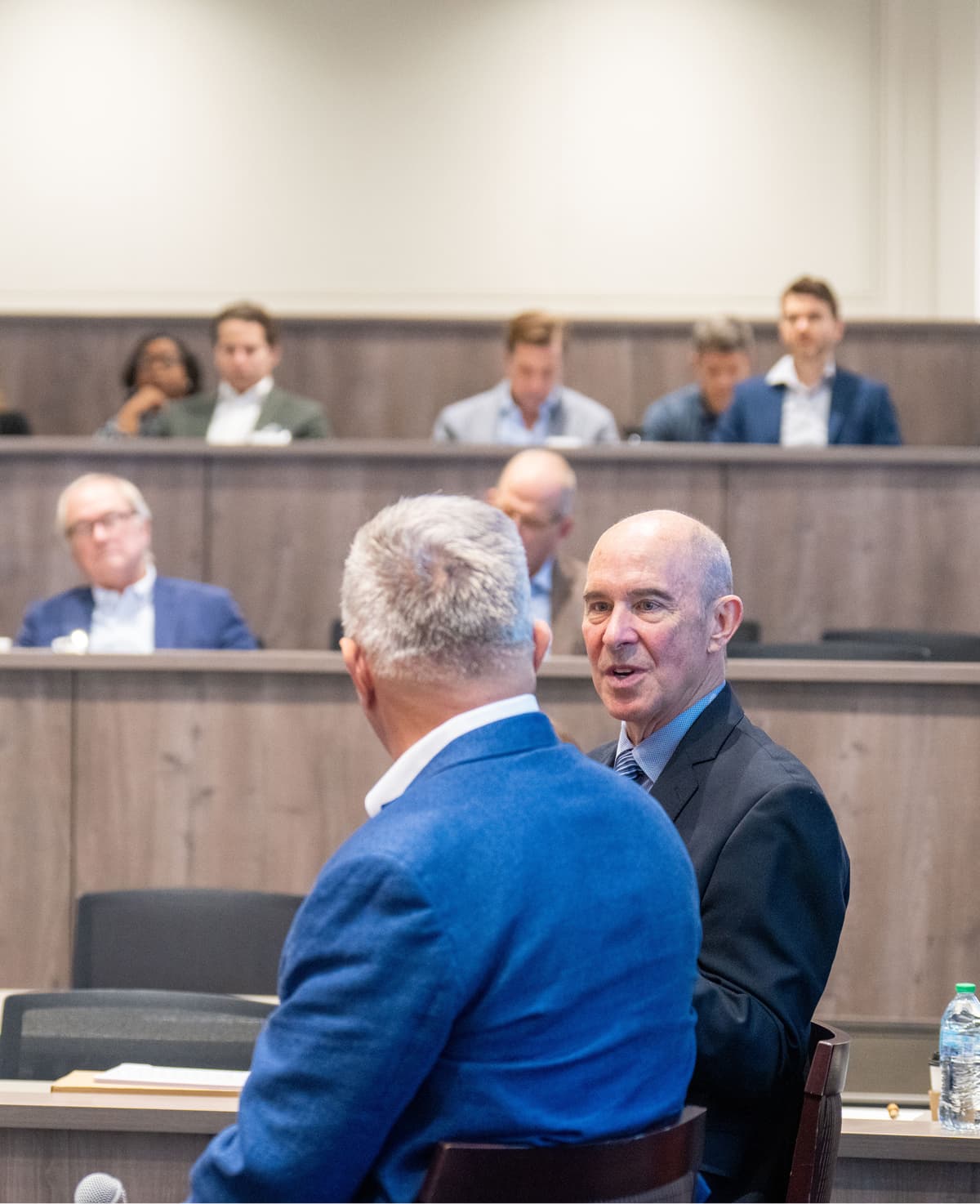 Two men in business attire talk before a session in a tiered lecture hall at Belmont University's Frist College of Medicine, with attendees seated behind them