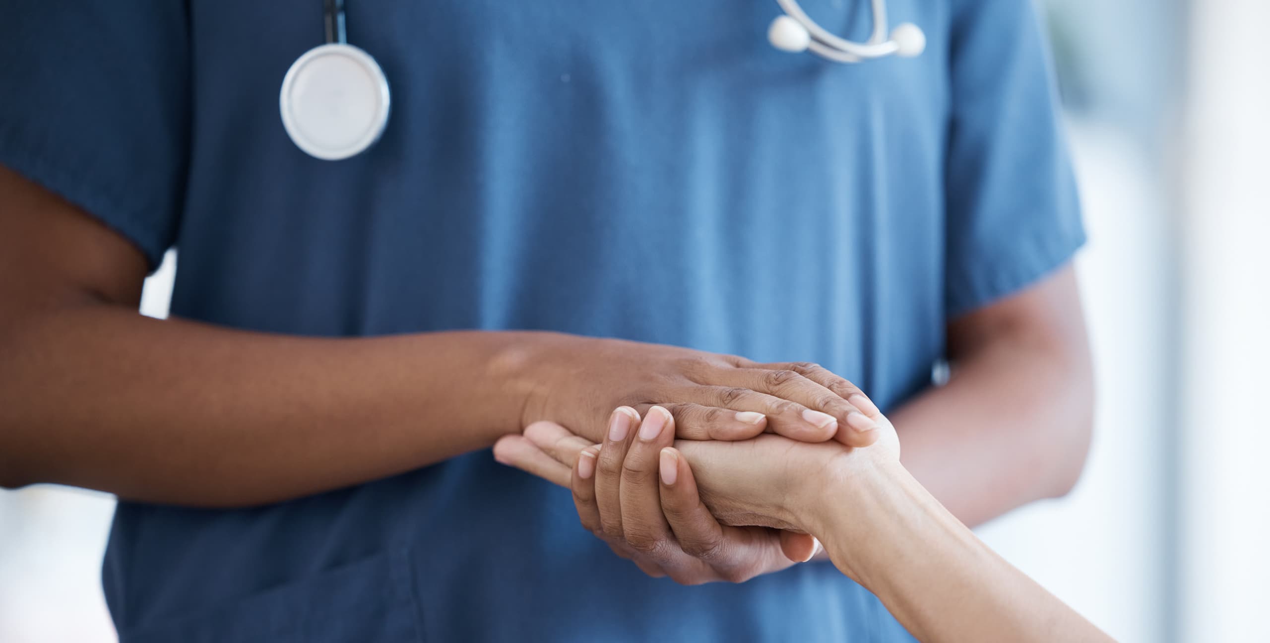 Close-up of a healthcare worker in blue scrubs and a stethoscope gently holding a patient's hand in a caring gesture.